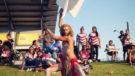 Aboriginal dancer in traditional costume waves a big maroon and white flag at a football game.