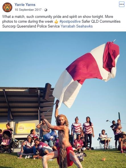 Aboriginal dancer in traditional costume waves a big maroon and white flag at a football game.