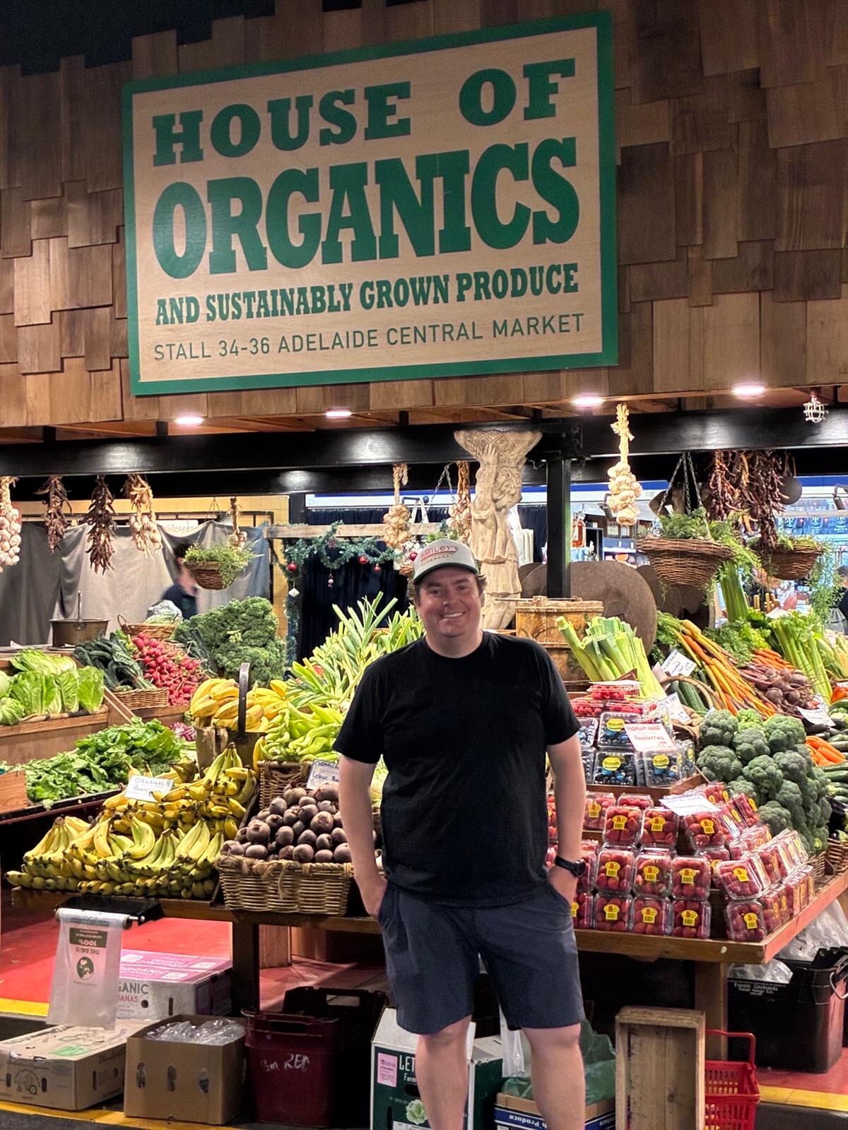 a man in cap, shorts and black t-shirt hands in pockets smiling in front of fruit and veg stall