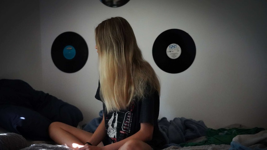 An unidentified teenage girl sitting on a bed with her legs cross and hair obscuring her face, with vinyl records on the wall.