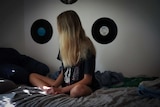 An unidentified teenage girl sitting on a bed with her legs cross and hair obscuring her face, with vinyl records on the wall.