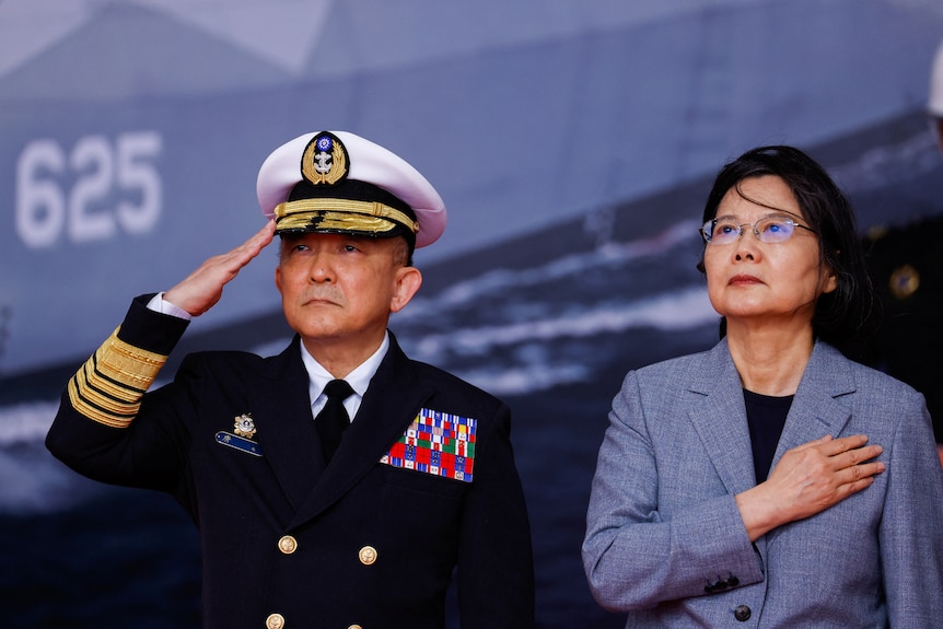 Taiwan President Tsai Ing-wen puts her right hand on her heart and she stands next to a saluting uniformed naval commander. 