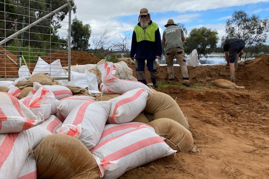 A pile of sandbags, behind them three men work to construct a levee bank.