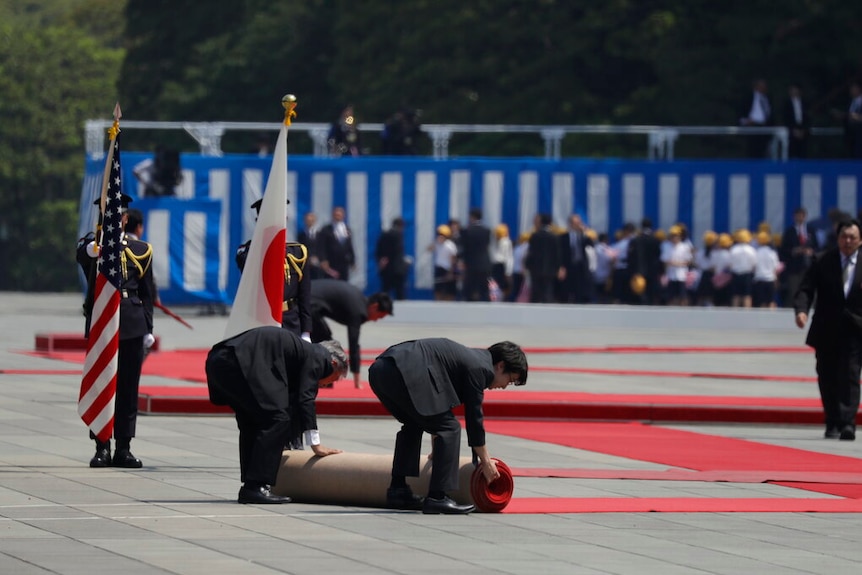 Men in suits roll up a red carpet while soldiers hold US and Japanese flags.