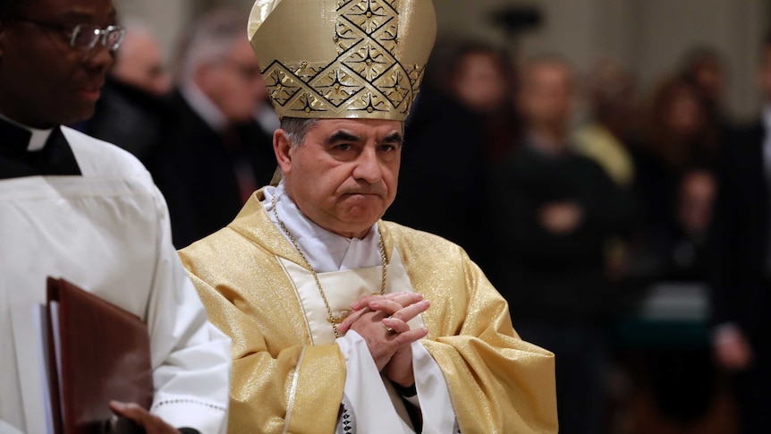 The cardinal wears gold robes and folds his hands in prayer.