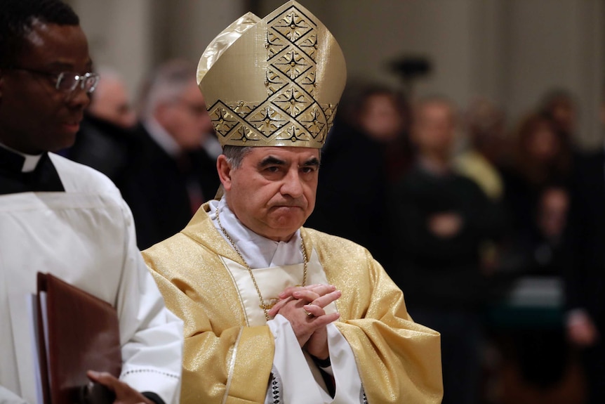 The cardinal wears gold robes and folds his hands in prayer.