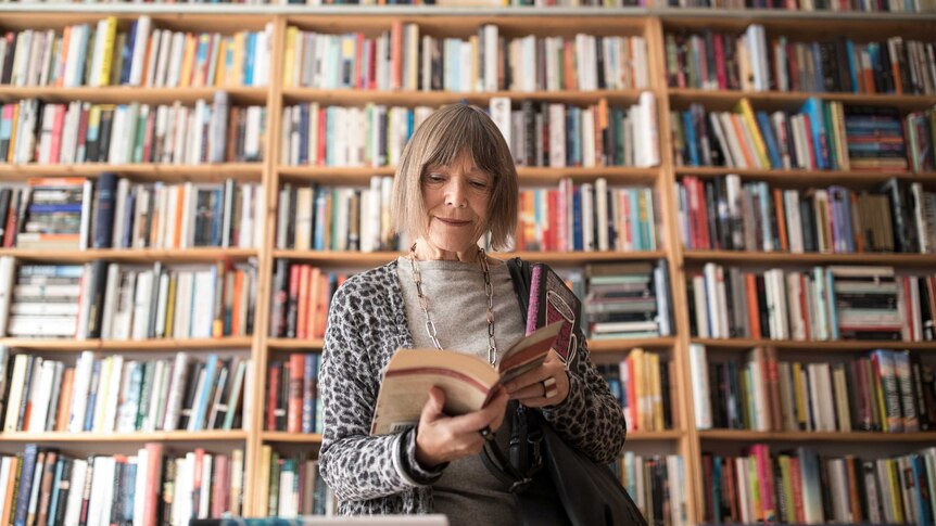 Woman with greyish hair smiles slightly while reading book. Behind her large floor-to-ceiling bookshelves are filled with books.
