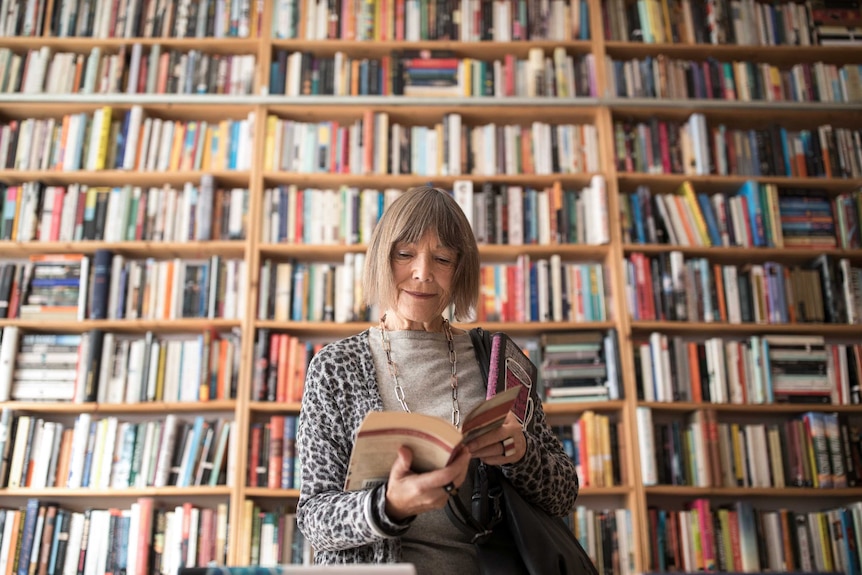 Woman with greyish hair smiles slightly while reading book. Behind her large floor-to-ceiling bookshelves are filled with books.