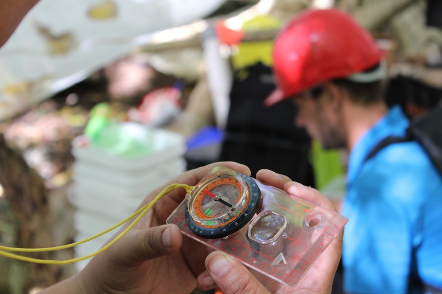 Hands hold a compass. In the background, a person wears a red hard hat.