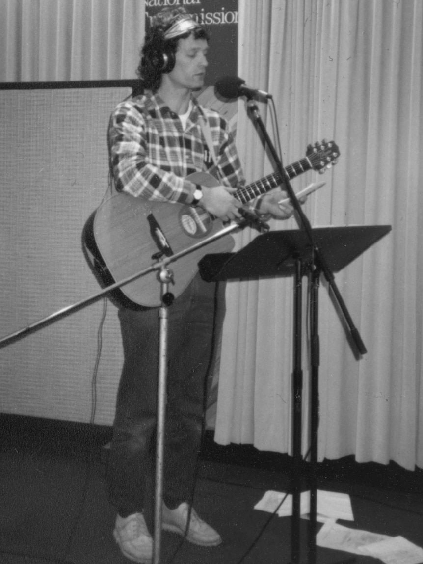Black and white photo of Champion holding guitar and standing in front of microphone in studio.