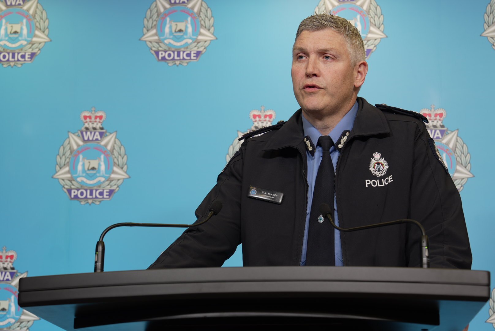 Coronel Blanch com uma jaqueta escura de policial falando aos repórteres atrás de um pódio na sede da polícia.