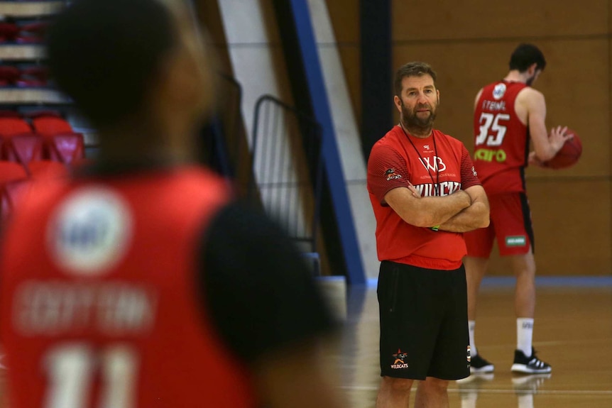 Perth Wildcats coach Trevor Gleeson stands with arms folded at training with a player in the foreground and another behind him.