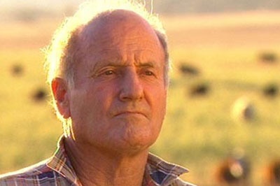 A close-up of a grey-haired man in a rural setting, with sheep visible in the background