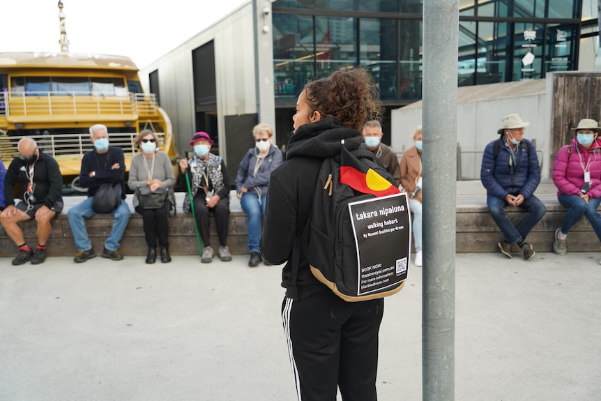 A woman wearing a backpack with a small Aboriginal flag sticking out of it has her back to the camera as she speaks to a group