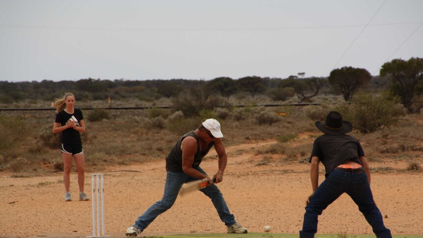 A man in a white cap and singlet stands on a cricket pitch ready to hit the ball with a wooden bat.