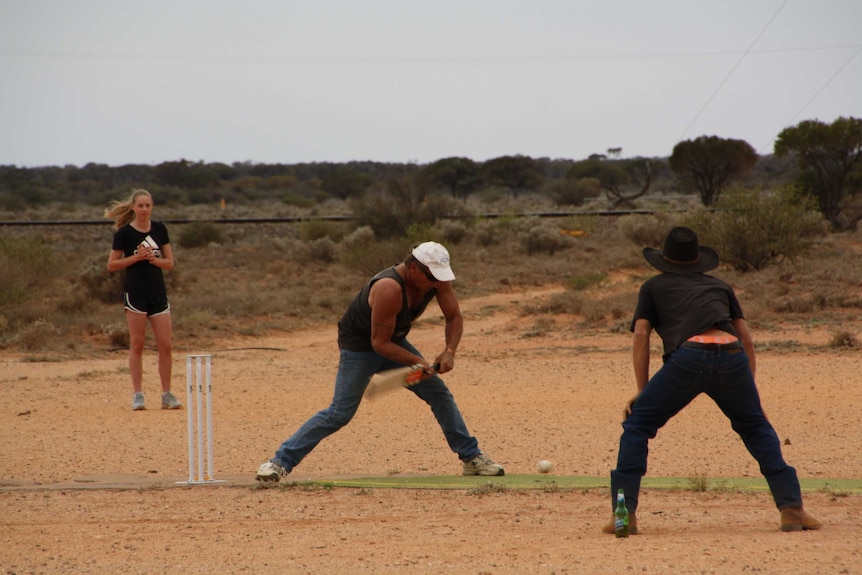 A man in a white cap and singlet stands on a cricket pitch ready to hit the ball with a wooden bat.