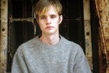A young man with brown hair, with blonde highlight at the tips, and a grey knit looking at the camera.