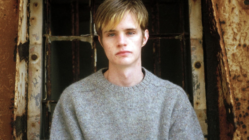 A young man with brown hair, with blonde highlight at the tips, and a grey knit looking at the camera.