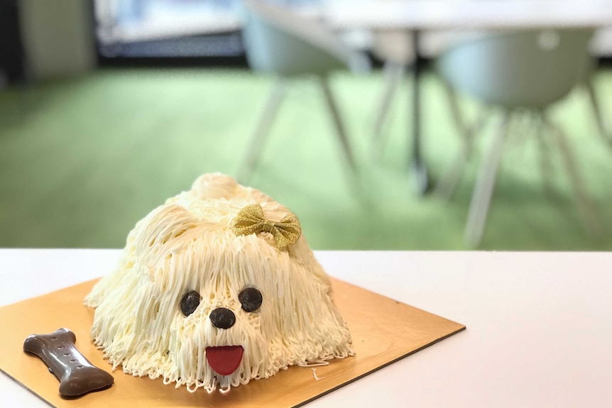 A white dog shape cake on the table where was with a dinning table behind.