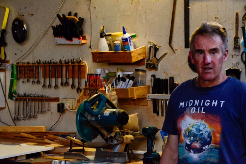 A man in a midnight oil shirt standing in front of a wall full of tools