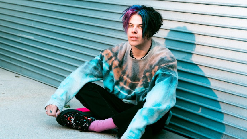 Press shot of Yungblud, sitting on ground against a garage door; dressed in black jeans and blue tie dye jumper