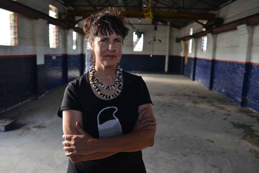 The artist Judy Watson standing in an empty factory space, looking determinedly at the camera