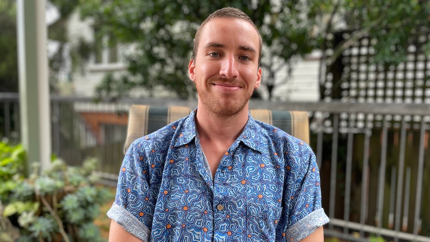 A man sits in an outdoor chair, wearing a patterned short-sleeved button-down shirt.