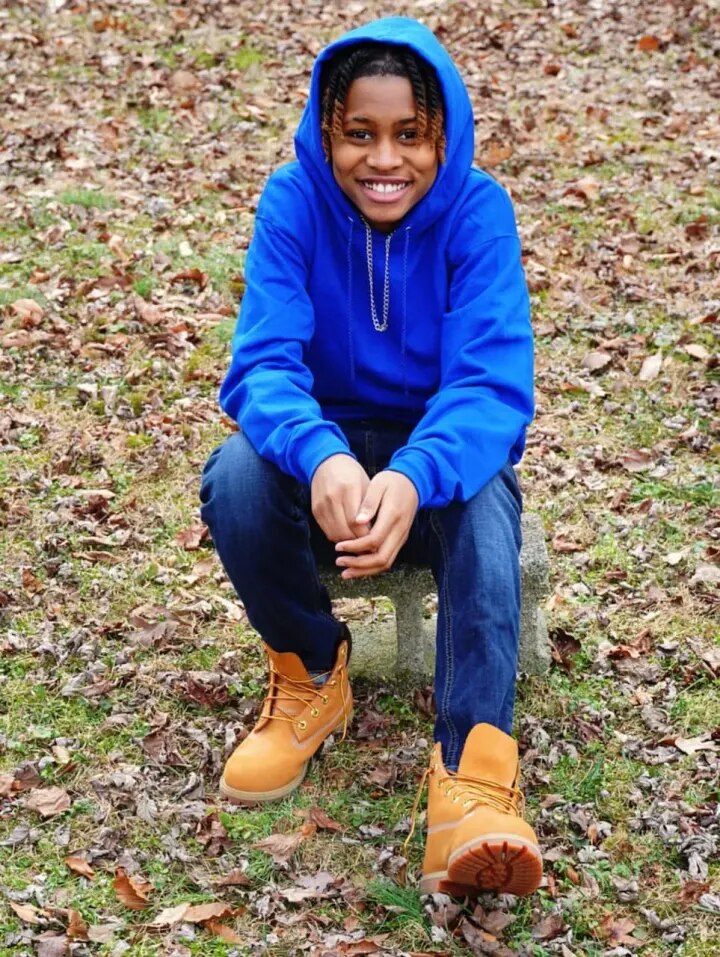 Naima wears a blue hoodie and jeans as she sits on a small chair on a lawn covered with autumn leaves.