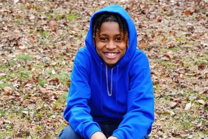Naima wears a blue hoodie and jeans as she sits on a small chair on a lawn covered with autumn leaves.