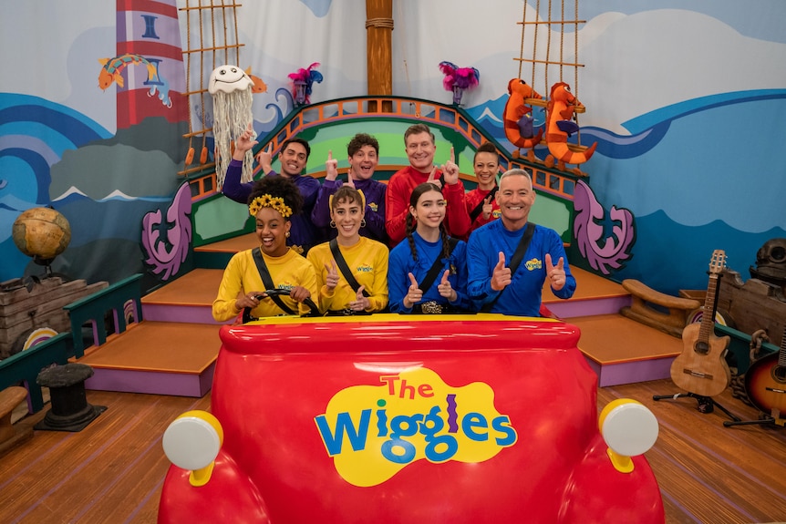 The eight people who now make up the Wiggles in purple, yellow, blue and red skivvys sitting inside a car with The Wiggles on it