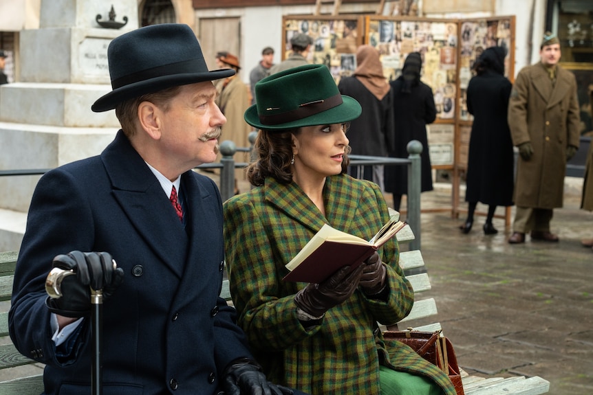 A film still showing a man and a woman in period dress sitting on a street bench, both wearing hats and suits and looking right