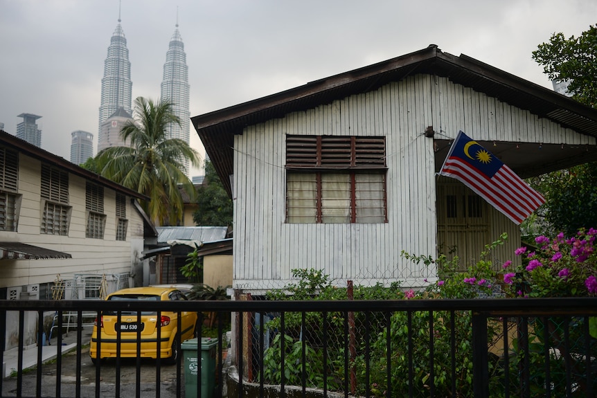 A Malaysian flag flies from an old, raised timber home. A yellow car is parked at the side. Skyscrapers are on the horizon.