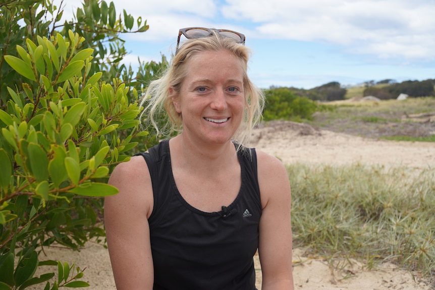 A woman sits on a sand dune and smiles at a camera.