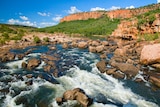 A river runs over rocks at the bottom of a rock red gorge with trees.