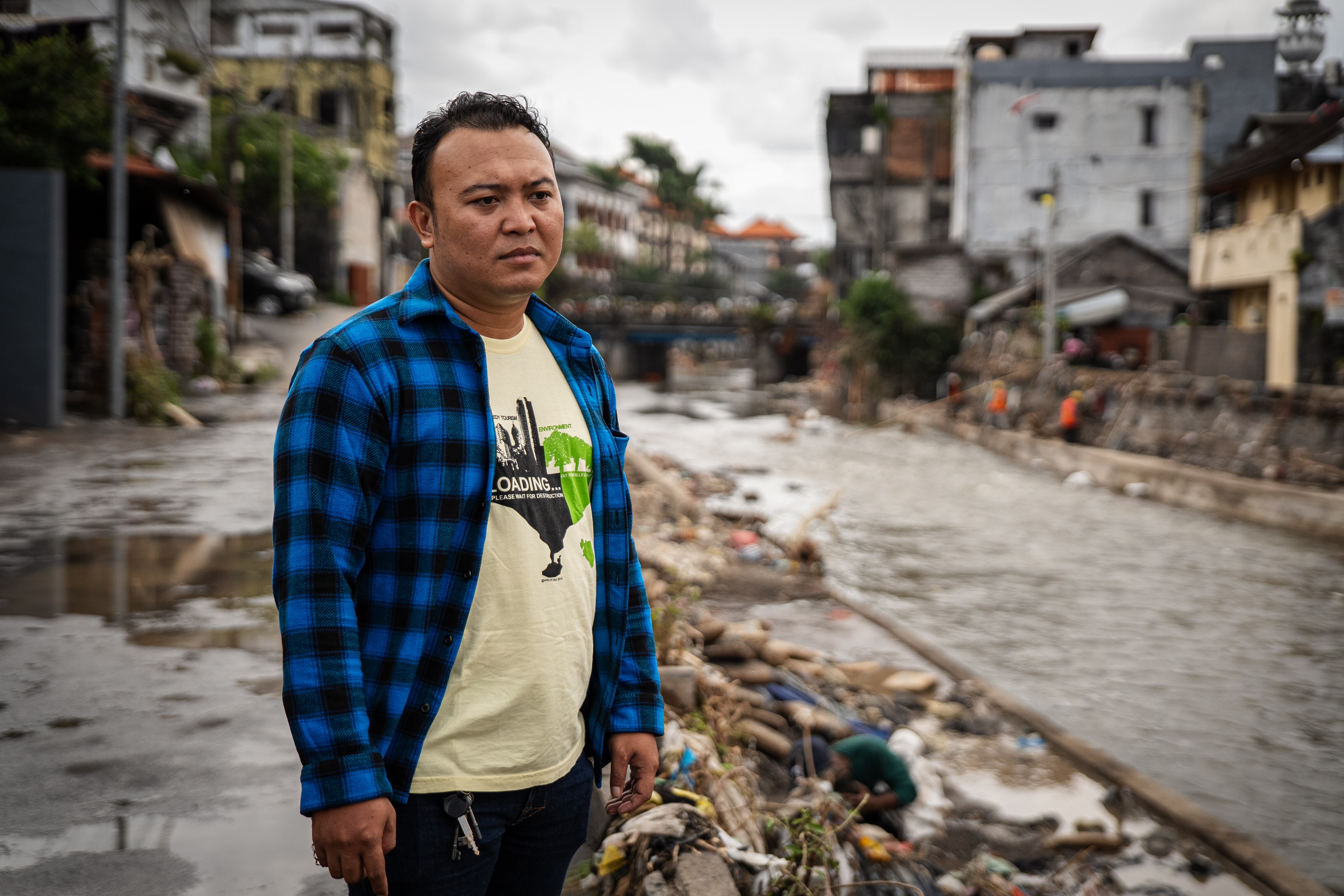 A man wearing a navy flannelette shirt over a tee stands beside a canal filled with trash