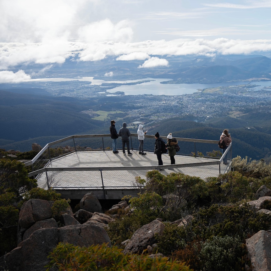 A wooden platform at the top of a mountain with people standing on it, looking out to spectacular view over a city