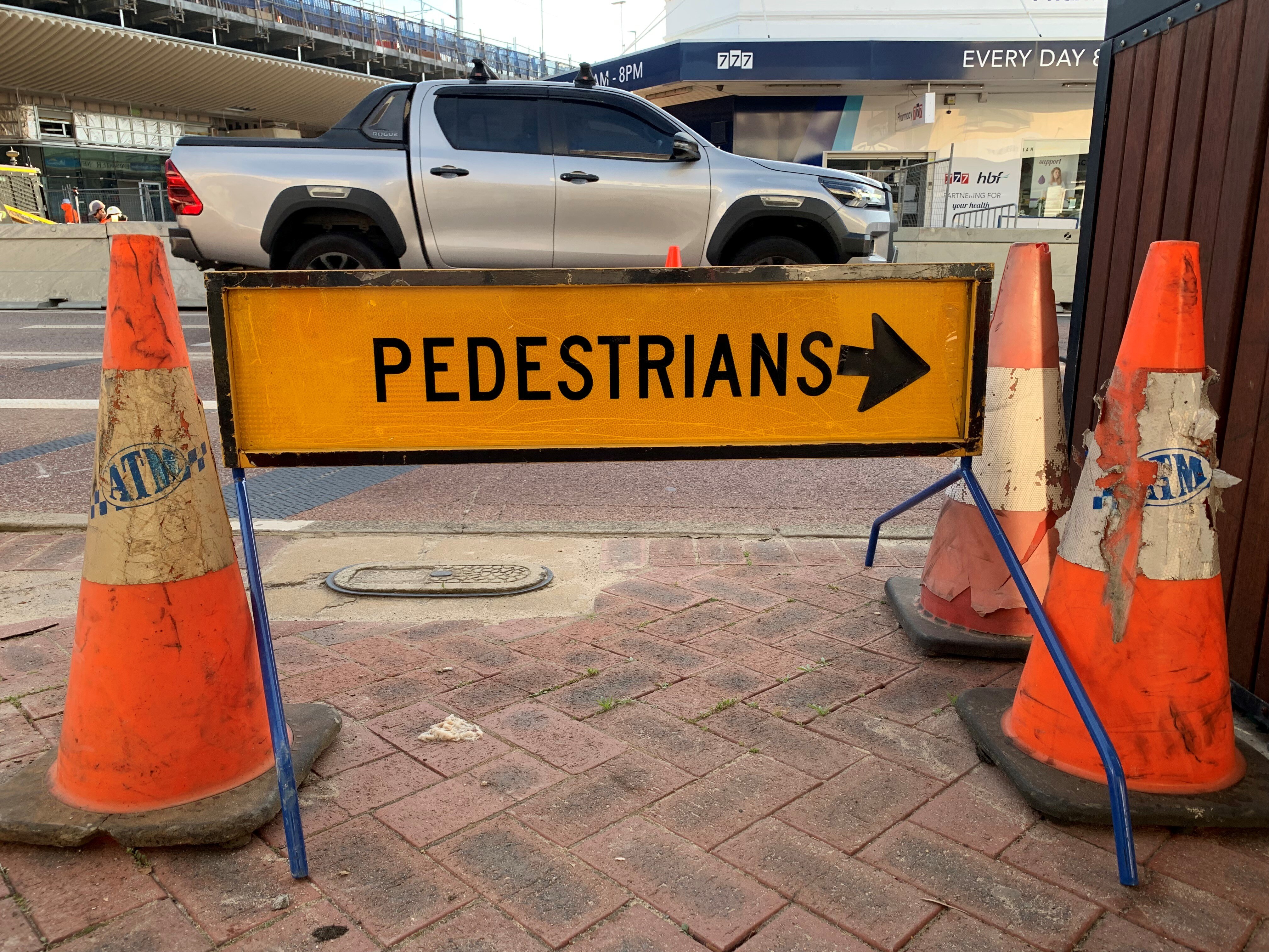 A yellow 'Pedestrians' sign with an arrow and three orange cones on a footpath in front of a yellow utility on the road.