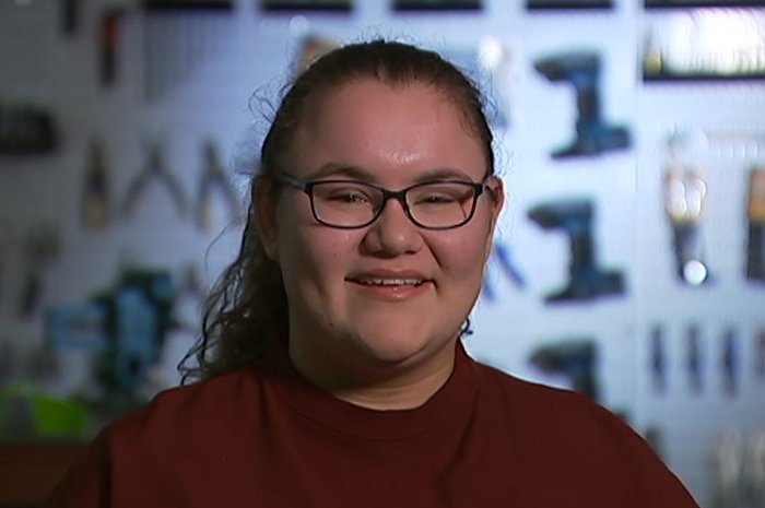 Katelinel Moores smiles at the camera, in front of a wall on which tools are hanging.