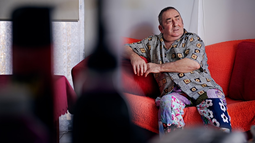 An older man sits on a red couch and looks away