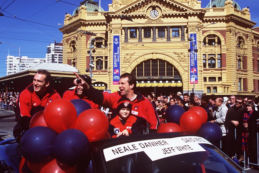 An AFL coach, two players and a child smile and point at the crowd as they ride in a car in the AFL grand final parade.
