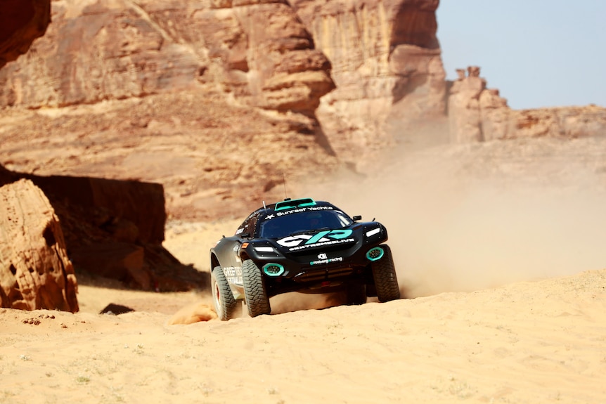A black car drives on sand with a rocky cliff behind it