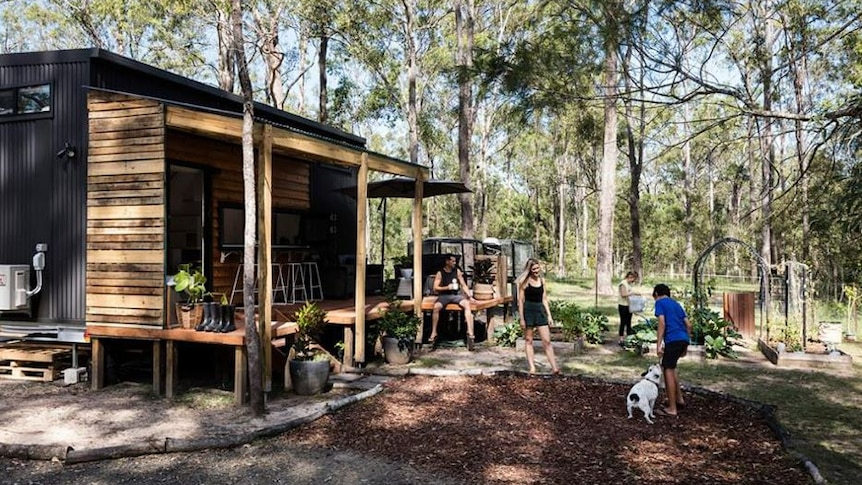 Boy and mother play with a dog in front of a tiny house surrounded by bushland. 