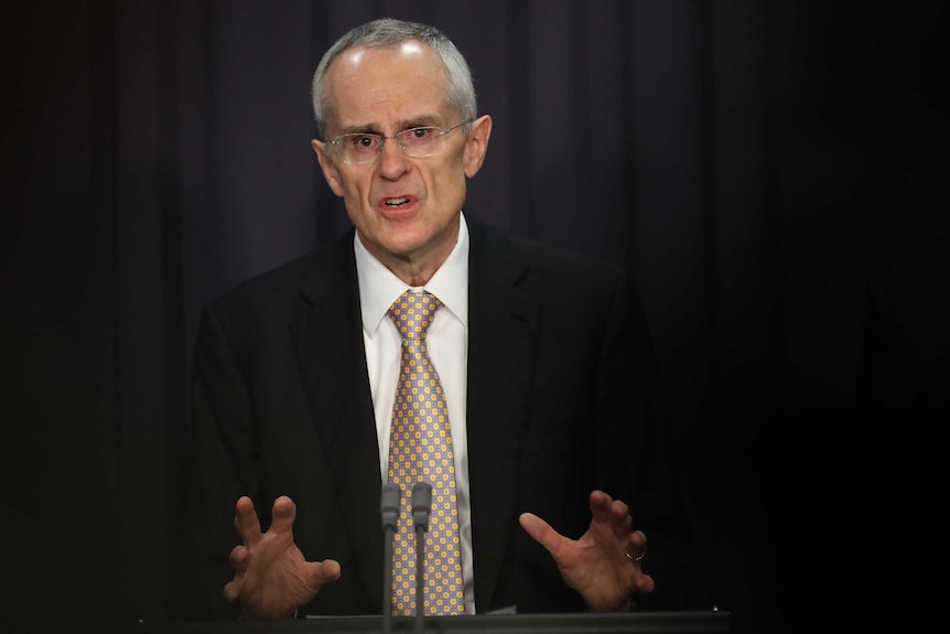 Chairman of the ACCC, Rod Sims addresses the media in the Blue Room. He's wearing glasses and an orange spotted tie.