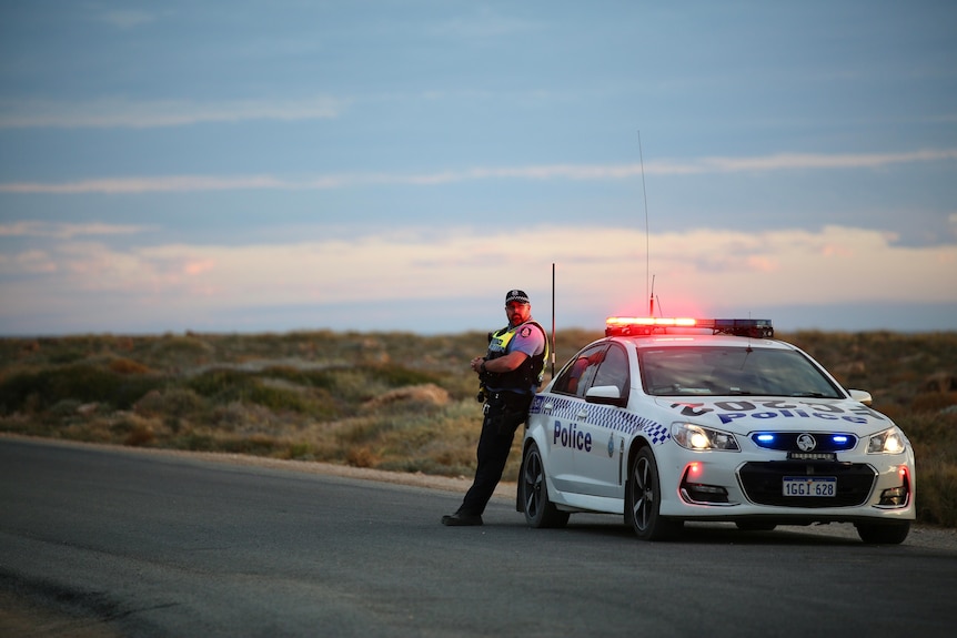 A police officer leans against a police car on a road in a isolated rugged coastal area at dusk.