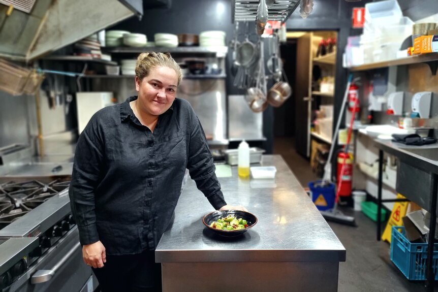 Woman dressed in black leans on stainless steel counter in commercial kitchen. Next to her is a freshly cooked plate of sprouts.