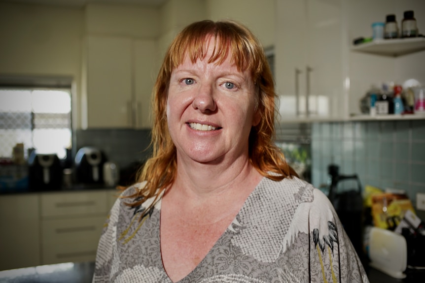 A woman with red hair smiles at the camera inside her kitchen at home