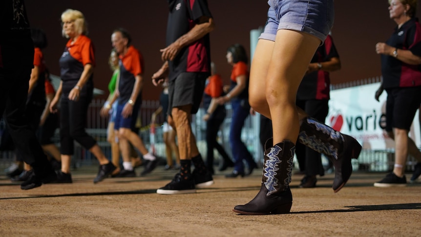 A group of people line-dancing in an outdoors area at dusk, with a woman's legs wearing cowboy boots in the foreground.