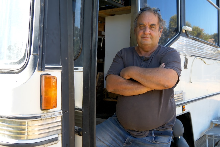 A man stands with his arms crossed in front of a bus. 