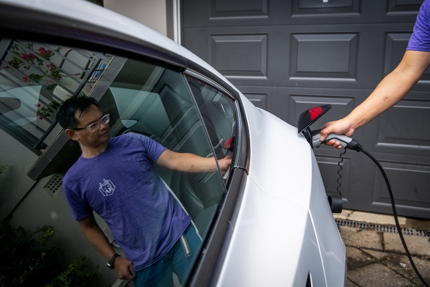 Marc Tan's reflection is seen in the shiny windows of his Tesla as he plugs the charger in.