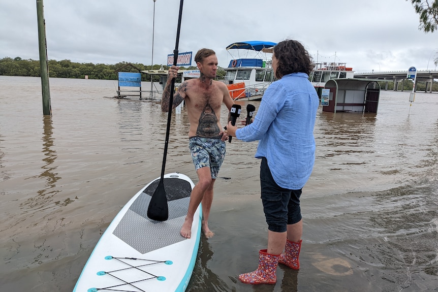 Journalist wearing gumboots interviewing man standing next to a paddle board in floodwaters.
